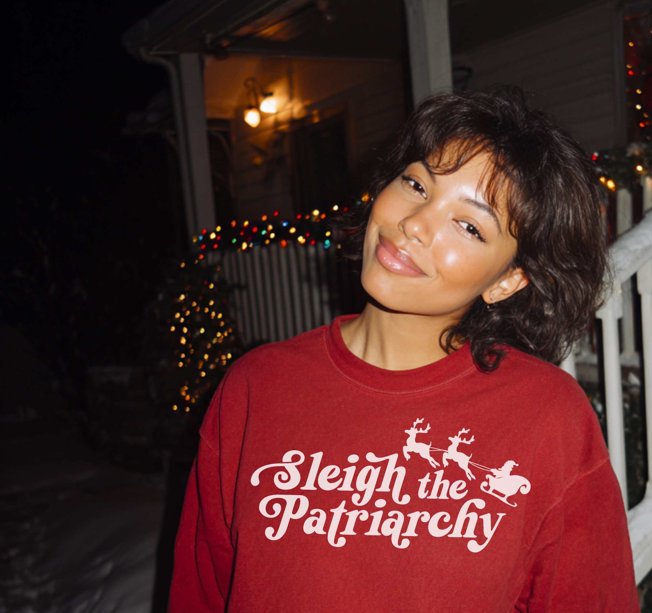 Person wearing a red sweater with 'Sleigh the Patriarchy' text, standing on a snowy porch.