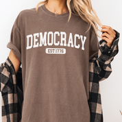 Person wearing a brown t-shirt with 'DEMOCRACY EST 1776' text.