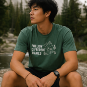 Man wearing a green t-shirt with 'Follow Different Trails' text and mountains graphic, sitting outdoors.